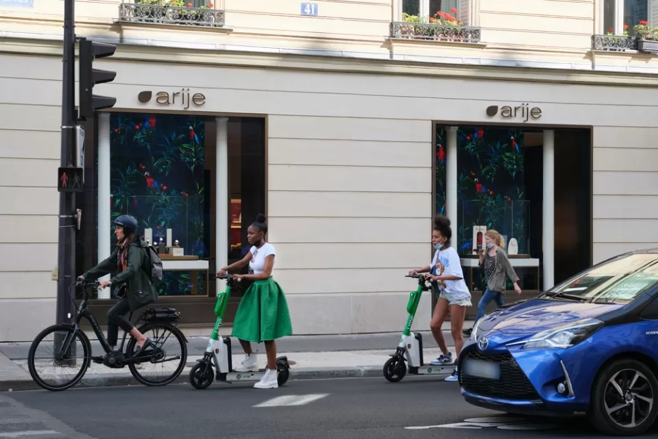 A woman on an electric bike and two women on electric scooters wait at an intersection for the light to change.